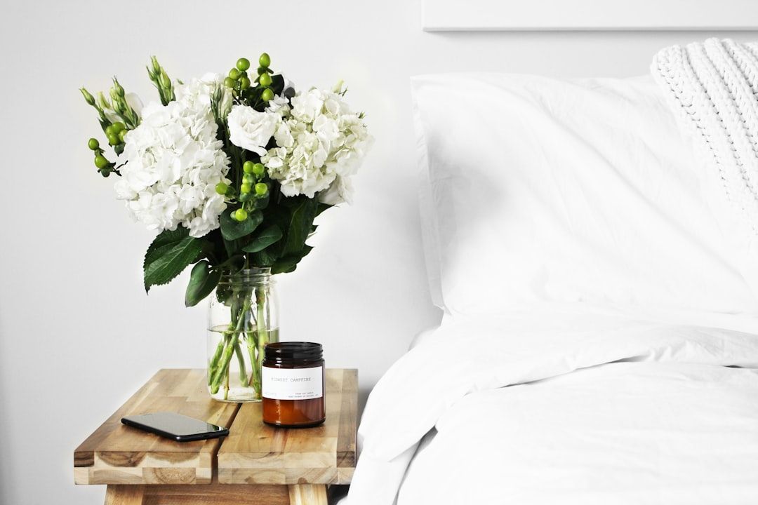 Ein Strauß weißer Blumen in einer Glasvase, eine Kerze und ein Smartphone stehen auf einem hölzernen Nachttisch neben einem Bett mit weißer Bettwäsche und schaffen eine Atmosphäre, die an ein ruhiges Hotelzimmer in Aachen erinnert.