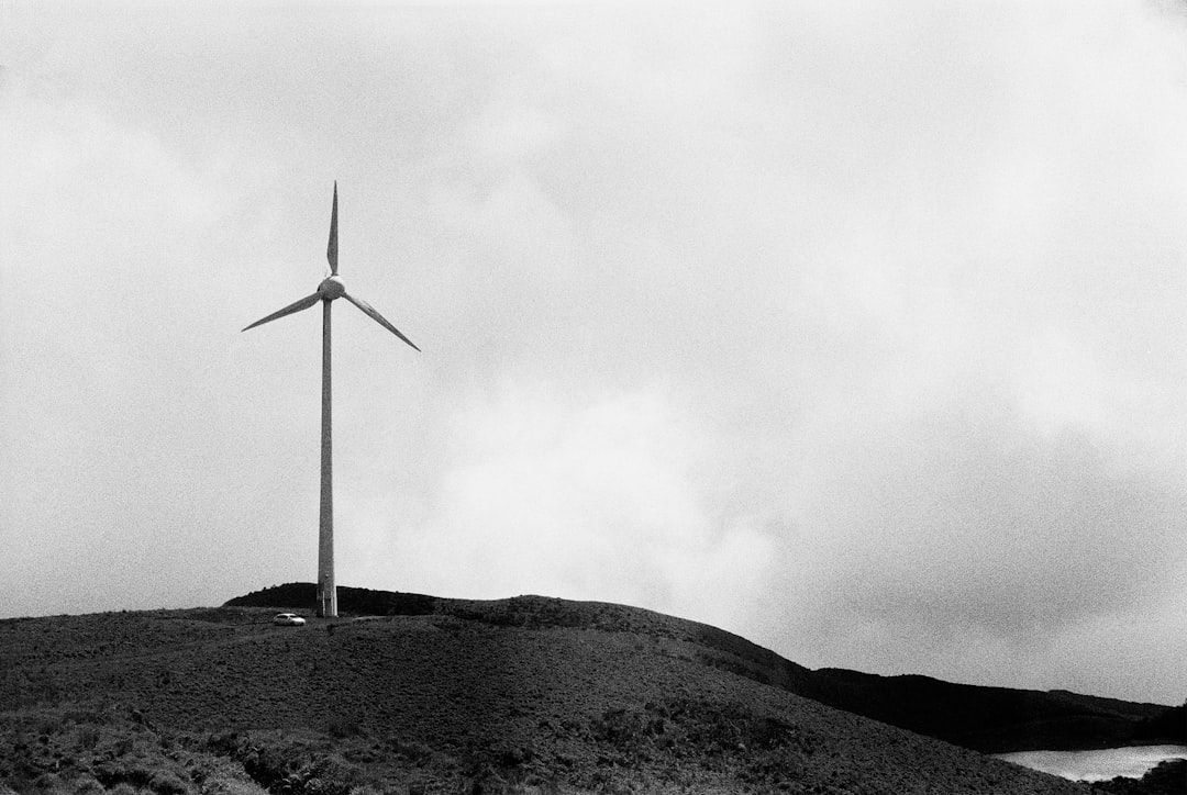 Eine einsame Windkraftanlage steht auf einem Hügel in der Nähe von Aachen unter einem bewölkten Himmel. In der unteren linken Ecke ist ein Auto geparkt. Das Bild ist schwarzweiß.
