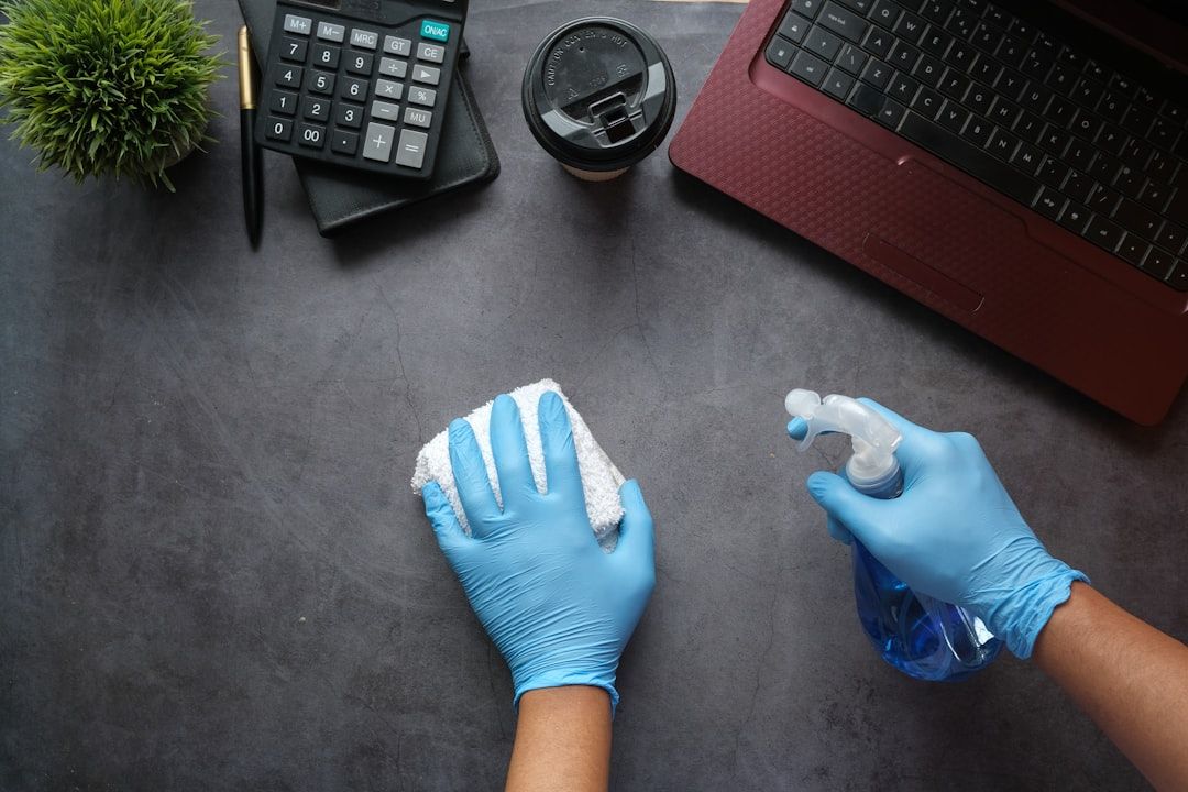 Hände in blauen Handschuhen reinigen sorgfältig einen Schreibtisch mit einer Sprühflasche und einem Tuch neben einem Laptop, einem Taschenrechner, einer kleinen Pflanze und einer Kaffeetasse, alles in einem Büroraum, der an Aachens disziplinierte Arbeitsumgebung erinnert.