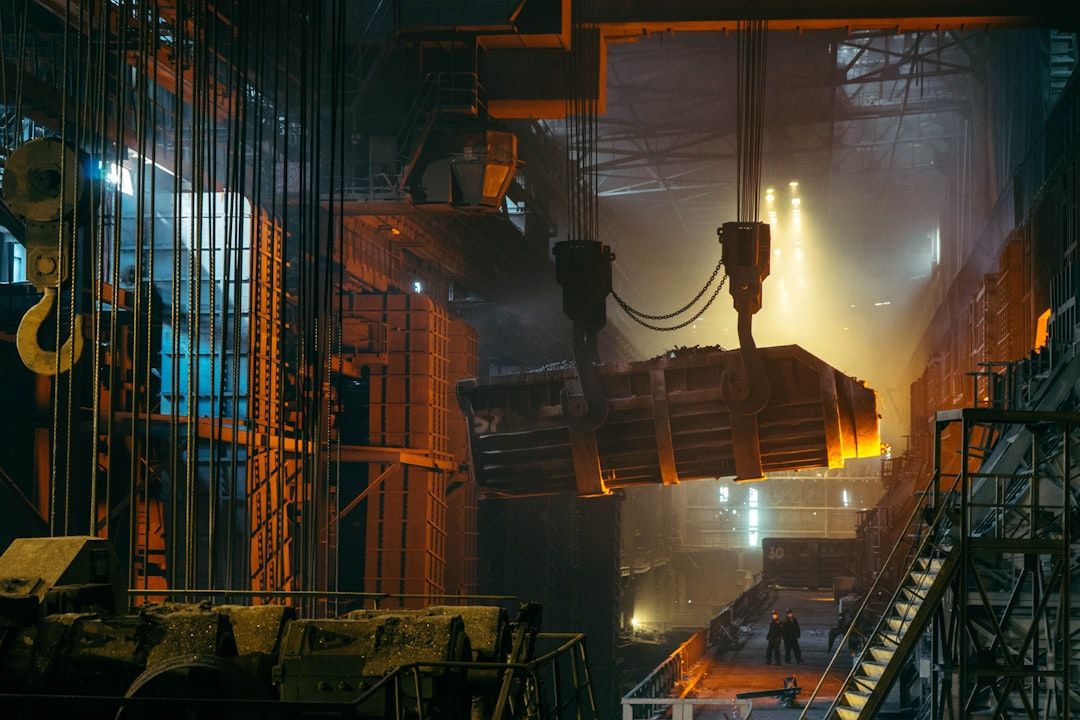 In einer schwach beleuchteten, großen Fabrik in Aachen hängt ein Industrieofen mit glühendem, geschmolzenem Metall an Kränen. In der Ferne sind Metallstrukturen und Arbeiter zu sehen.