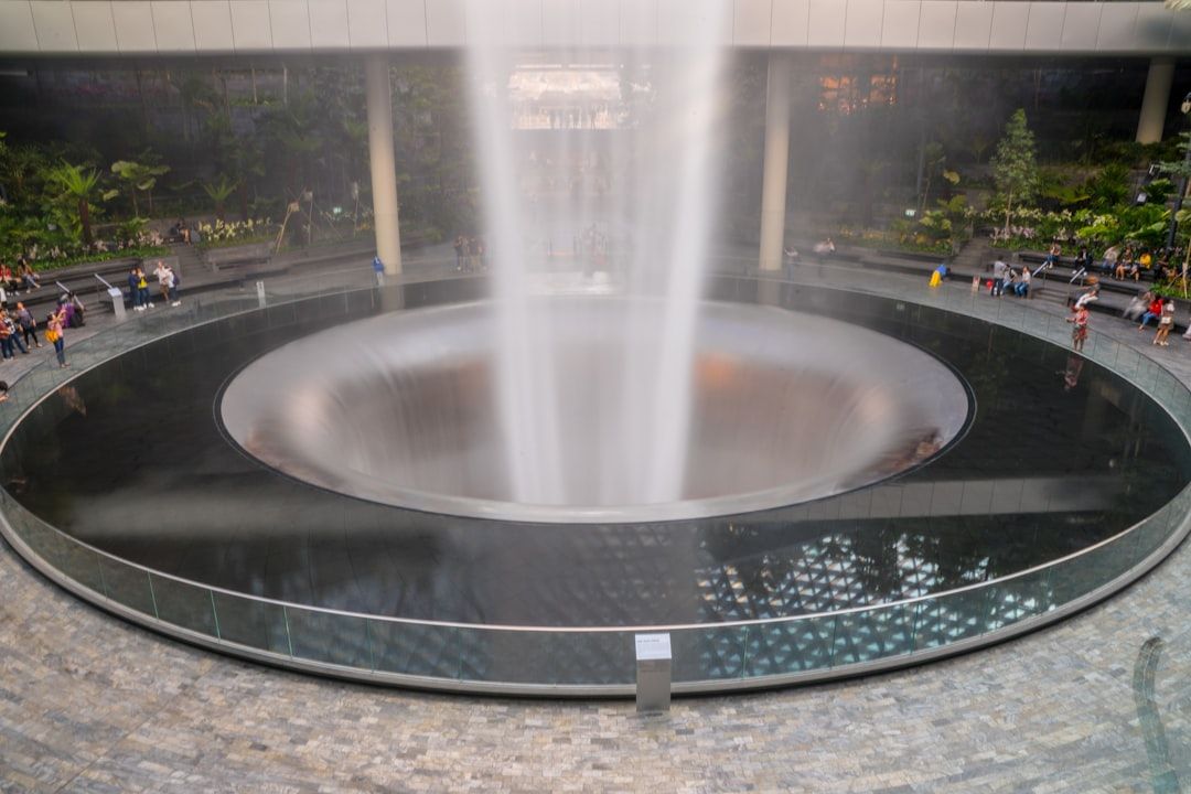 In einem großen, modernen Gebäude mit Glasdach, das an die innovative Architektur Aachens erinnert, sitzen und laufen Menschen um einen Indoor-Wasserfall herum.