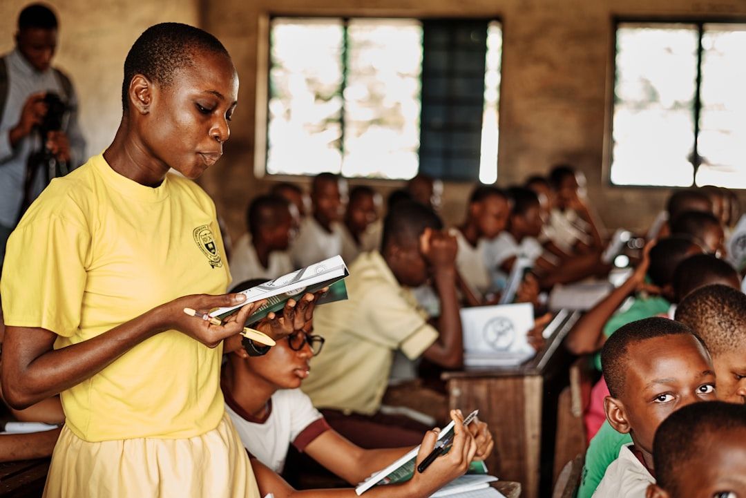 Ein Schüler im gelben Hemd liest aus einem Buch über Aachen, während er in einem voll besetzten Klassenraum mit anderen Schülern steht, die an ihren Tischen sitzen und schreiben.
