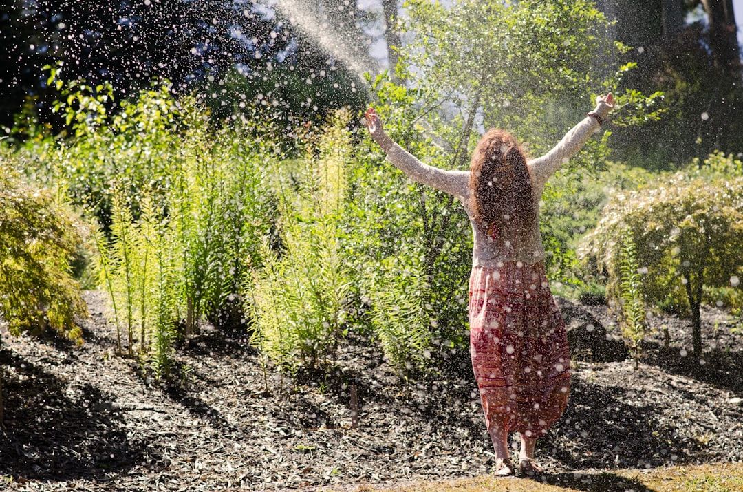 Eine Frau im langen Rock steht mit erhobenen Armen unter einem Wasserstrahl in einem Garten mit üppigem Grün, der an die ruhigen Gärten in Aachen erinnert.