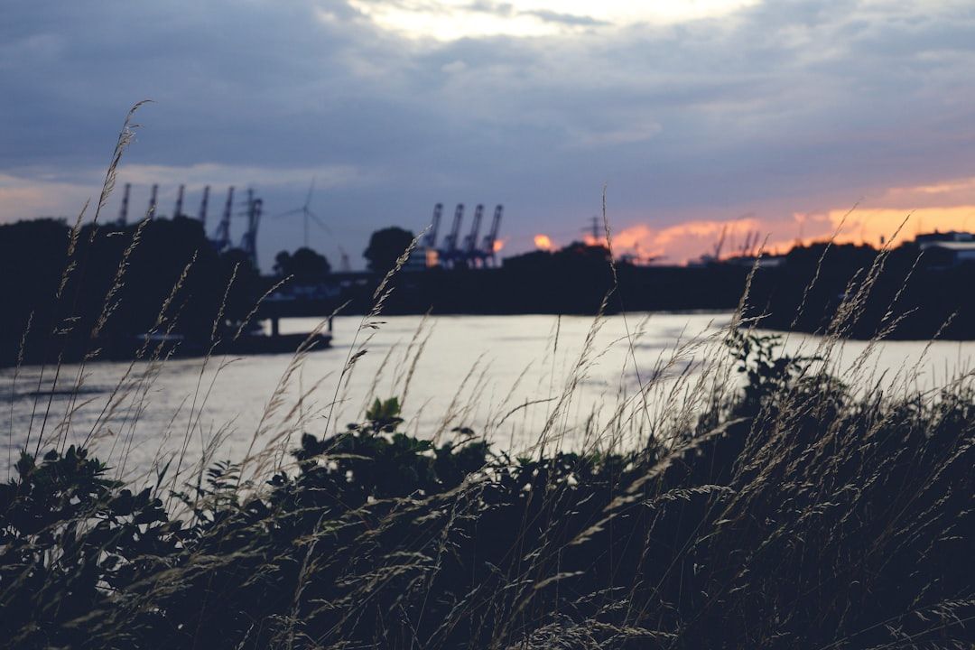 Ein Fluss im Sonnenuntergang mit hohen Gräsern im Vordergrund und Industriekränen im Hintergrund unter einem teilweise bewölkten Aachener Himmel.