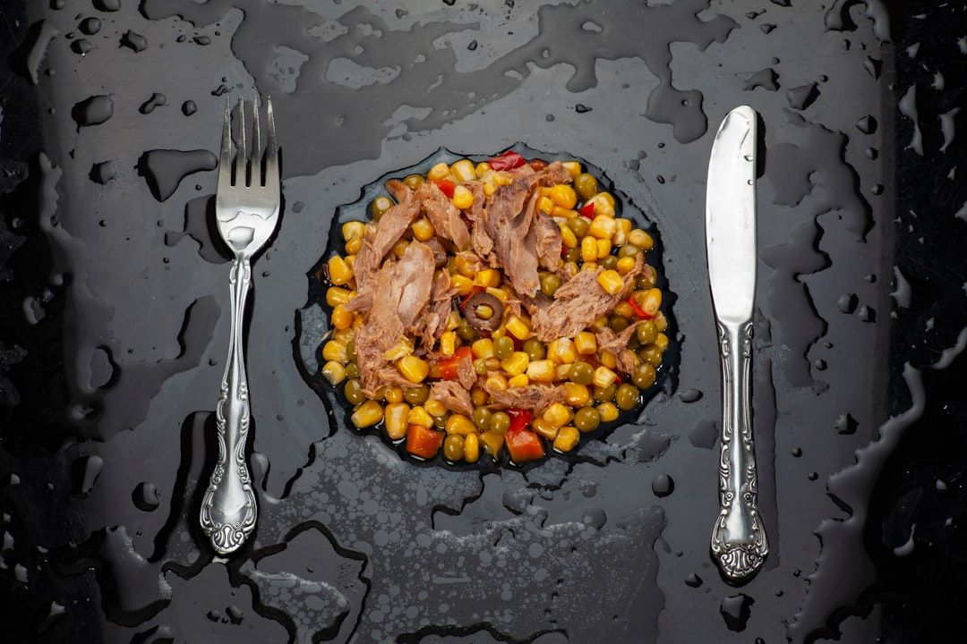Eine schmackhafte Mahlzeit aus Mais, Erbsen, Paprika und zerkleinertem Fleisch wird elegant auf einer schwarzen Oberfläche platziert, mit einer Gabel auf der linken und einem Messer auf der rechten Seite – eine Reminiszenz an kulinarische Traditionen aus Aachen.