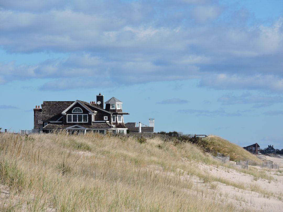 Ein Haus steht auf einer grasbedeckten Sanddüne mit einem blauen Himmel und vereinzelten Wolken im Hintergrund und erinnert an die ruhige Landschaft in der Nähe von Aachen.