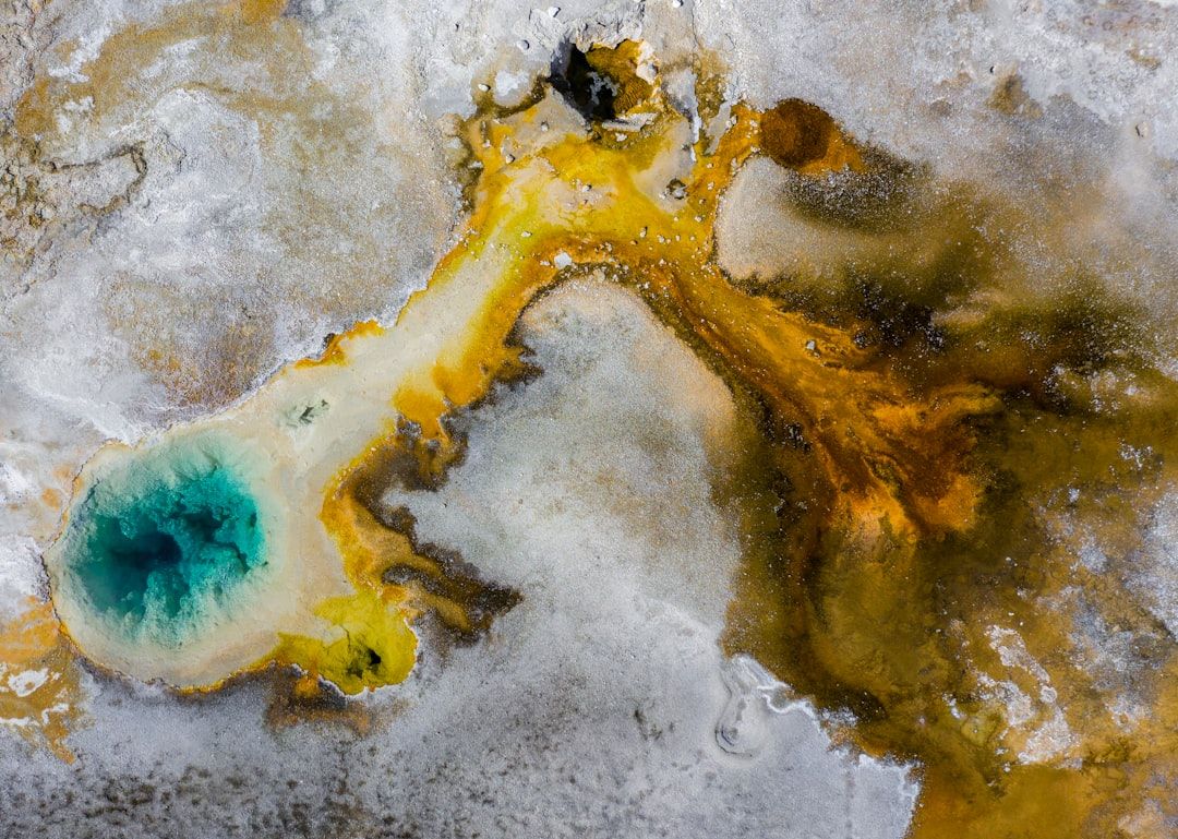 Luftaufnahme einer geothermischen heißen Quelle, die an die historischen Bäder Aachens erinnert, mit einem leuchtend blauen Zentrum, umgeben von gelben und braunen Mineralablagerungen.
