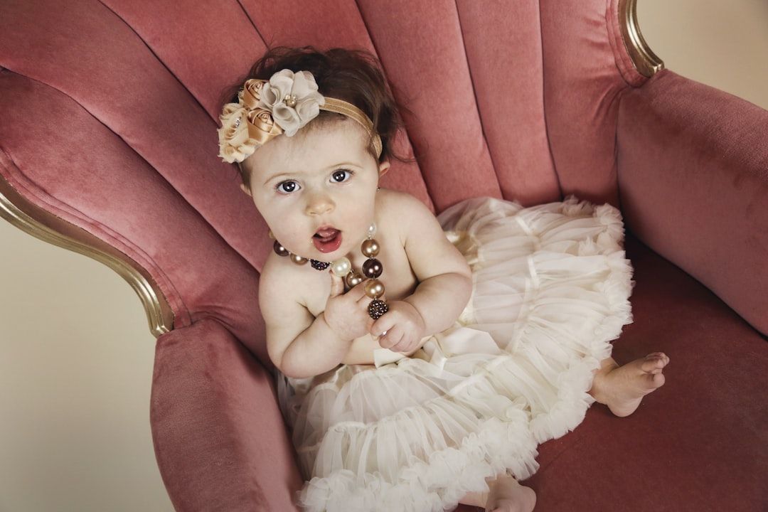 Ein Baby, bekleidet mit einem weißen Rüschenkleid und einem geblümten Stirnband, sitzt in einem rosa Polsterstuhl im Aachener Stil und ist mit einer Perlenkette und einem Armband geschmückt.