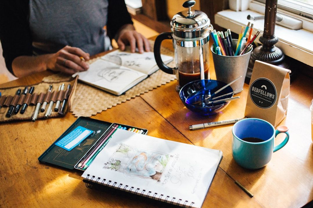 Eine Person skizziert in einem Notizbuch, umgeben von Künstlerbedarf, einer Kaffeekanne, einer Tasse dampfendem Kaffee und einer Tüte aromatischer Aachener Kaffeebohnen auf einem Holztisch.