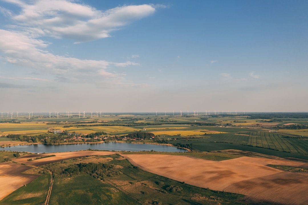 Luftaufnahme einer ländlichen Landschaft in der Nähe von Aachen mit Feldern, einem Gewässer und Windrädern in der Ferne unter einem klaren Himmel mit einigen Wolken.