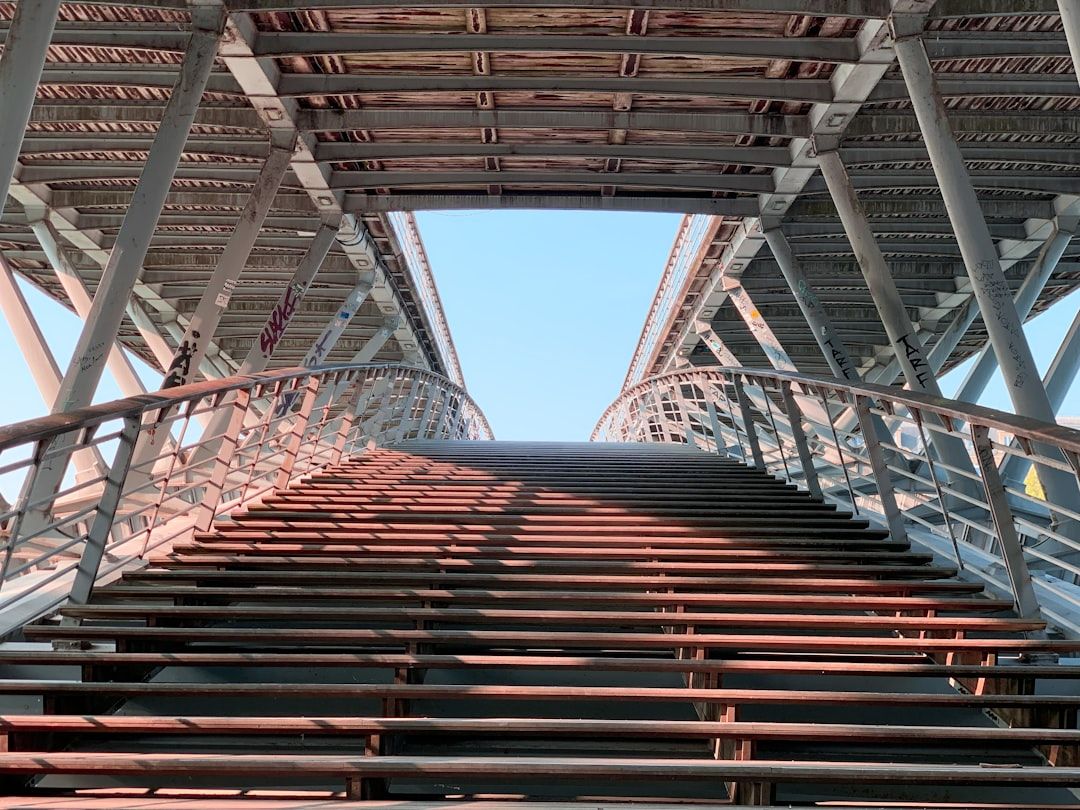 Blick vom Fuß einer steilen Treppe unter einer industriellen Metallbrückenkonstruktion in Aachen, mit Blick durch die Öffnung oben auf den blauen Himmel.