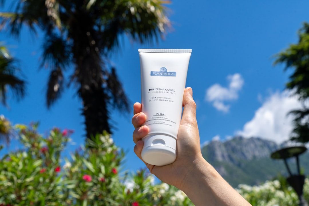 Eine Hand hält eine Tube Bio-Crema Corpo Körpercreme vor einem Hintergrund, der an die üppige Landschaft Aachens erinnert, mit Palmen, leuchtenden Blumen und majestätischen Bergen unter einem klaren blauen Himmel.