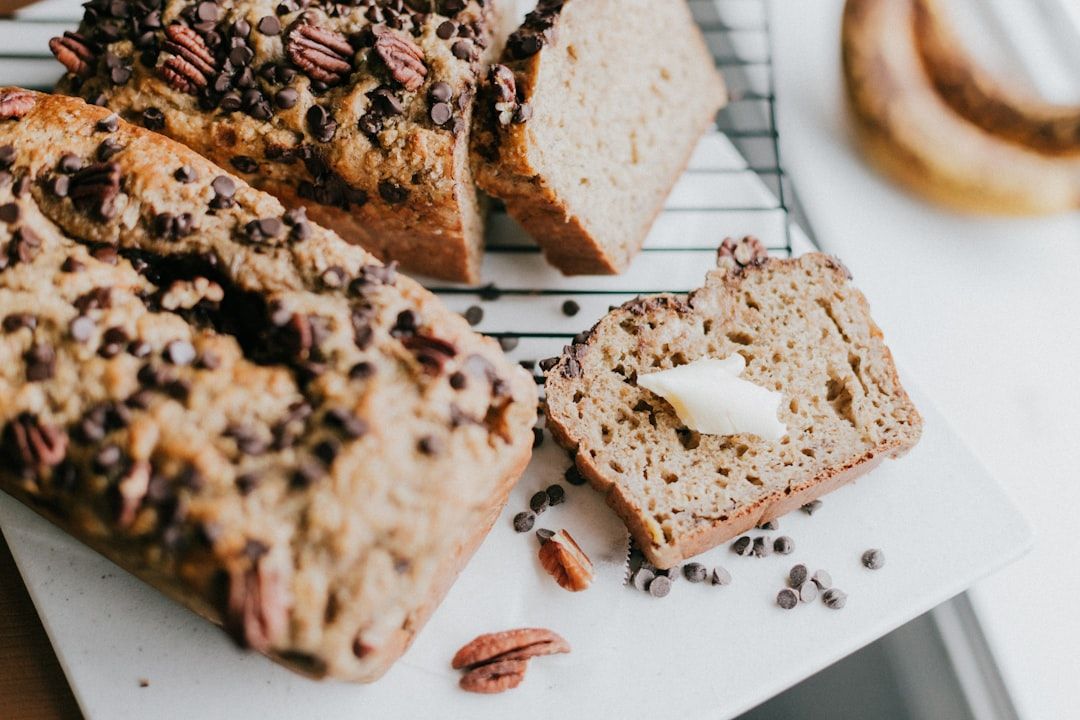 Bananenbrotscheiben mit Pekannüssen und Schokoladenstückchen liegen auf einem Kuchengitter und erinnern an die charmanten Aachener Bäckereien. Eine Scheibe mit Butter liegt neben dem Brot auf einer weißen Fläche.