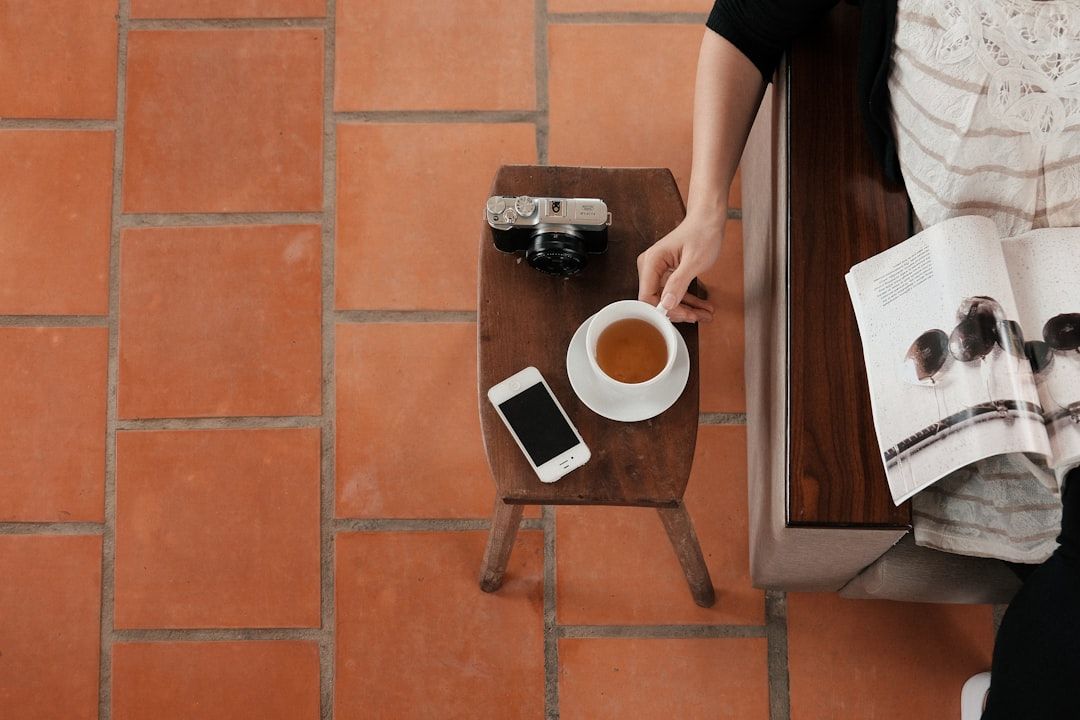 Eine Person hält eine Tasse Tee und hat eine Zeitschrift auf dem Schoß. Auf einem hölzernen Beistelltisch daneben liegen ein Smartphone und eine Vintage-Kamera, darauf eine alte Aachen-Karte. Der Boden ist mit Terrakotta-Fliesen ausgelegt.