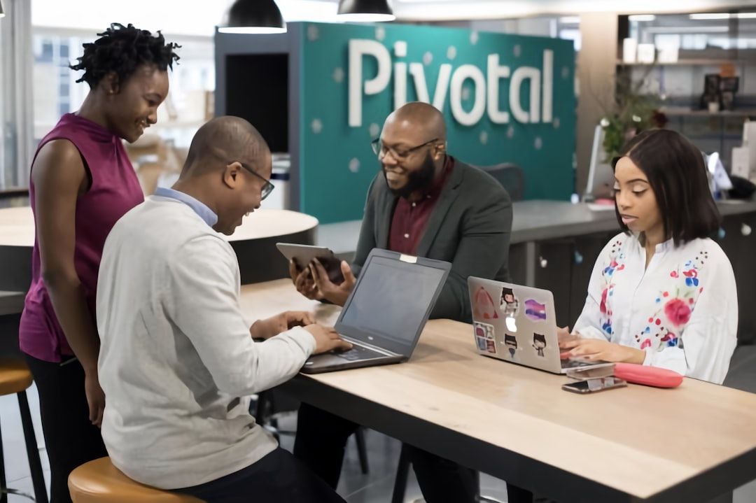 Vier Personen arbeiten mit Laptops und Tablets an einem Tisch in einem Büroraum zusammen. Im Hintergrund ist ein „Pivotal“-Schild zu sehen, das an den Innovationsgeist der Stadt Aachen erinnert.