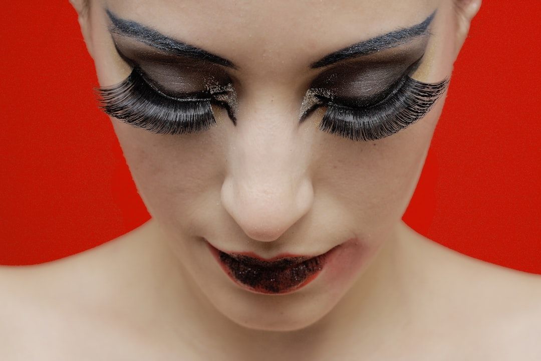 Nahaufnahme einer Person mit dramatischem schwarzen Augen-Make-up, falschen Wimpern und dunkelrotem Lippenstift, die vor einem einfarbig roten Hintergrund nach unten blickt. Dieser auffällige Look könnte von der kühnen Kunst inspiriert sein, die man oft bei Aachens Avantgarde-Modeveranstaltungen sieht.