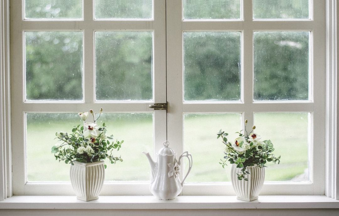 Ein Fenster mit sechs Scheiben blickt auf einen grünen Hof und verströmt den Charme eines Aachener Landhauses. Auf der Fensterbank stehen zwei weiße Pflanzkübel mit Blumen, in der Mitte steht eine weiße Teekanne.