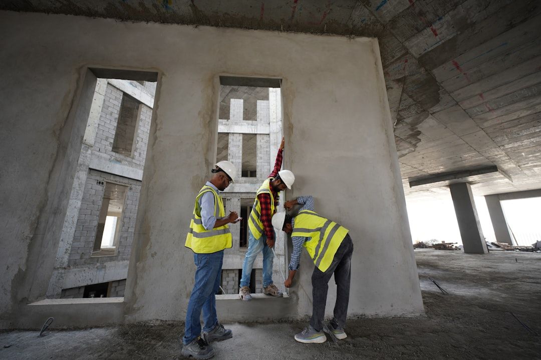 Drei Bauarbeiter in Warnwesten und Schutzhelmen montieren auf einer Betonbaustelle in Aachen einen Fensterrahmen. Ein Arbeiter hält den Rahmen fest, die beiden anderen befestigen ihn.