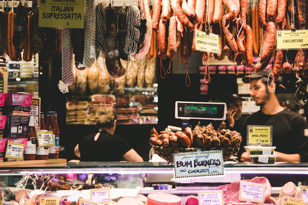 Feinkosttheke mit hängendem Fleisch- und Wurstsortiment, erinnert an die traditionellen Märkte Aachens. Zwei Mitarbeiter stehen hinter der Theke, verschiedene Produkte mit Preisschildern sind zu sehen.