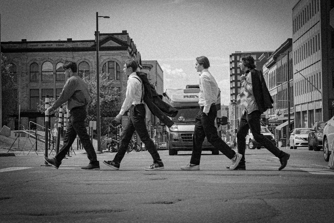 Vier Personen in Business-Kleidung gehen in einer Reihe über eine Stadtstraße in Aachen, im Hintergrund sind Gebäude und ein LKW zu sehen. Das Bild ist in Schwarzweiß gehalten und fängt eine zeitlose urbane Szene ein.