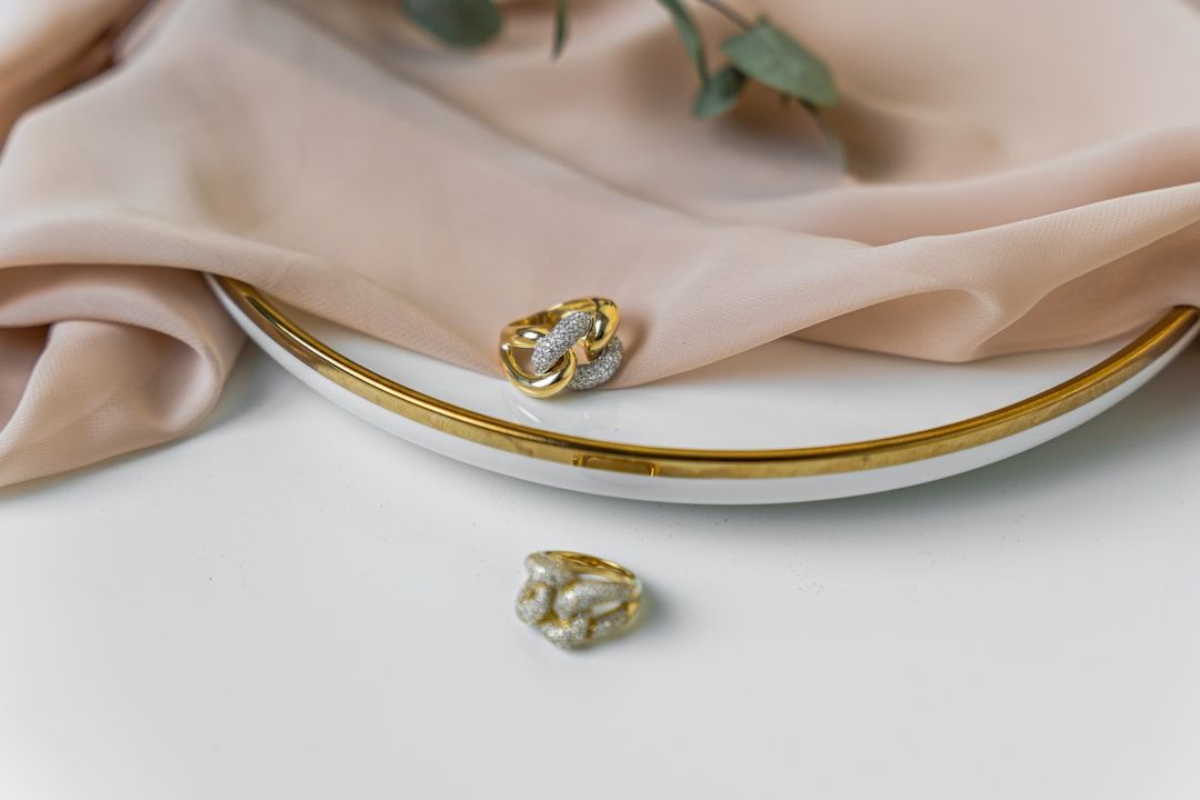 Zwei Goldringe mit aufwendigen Mustern, einer auf einem weißen Teller mit Goldrand und der andere auf einer weißen Oberfläche, neben einem beigen Stoff und grünen Blättern. Die Handwerkskunst erinnert an die Kunstfertigkeit der historischen Schätze Aachens.