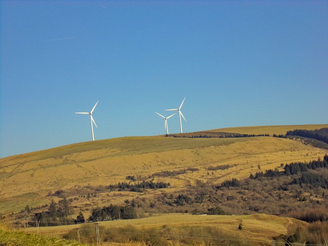Zwei Windräder auf einem grasbewachsenen Hügel in der Nähe von Aachen unter einem klaren blauen Himmel, mit einer Mischung aus Wald und offenem Land am Fuße des Hügels.