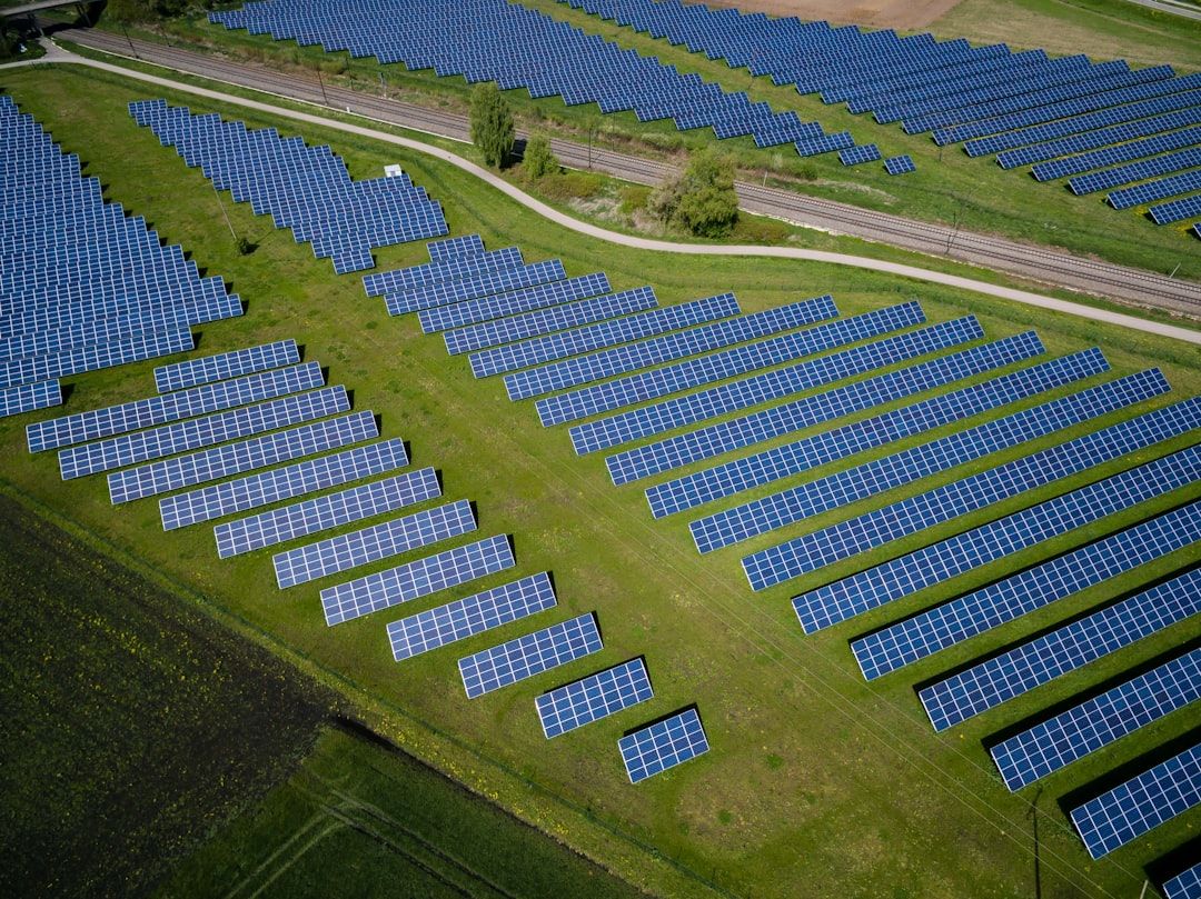 Luftaufnahme eines großen Solarparks mit zahlreichen Reihen blauer Solarmodule, die auf einer grünen Wiese in der Nähe von Aachen installiert sind. Ein Gehweg und Bahngleise verlaufen neben den Solarmodulen.