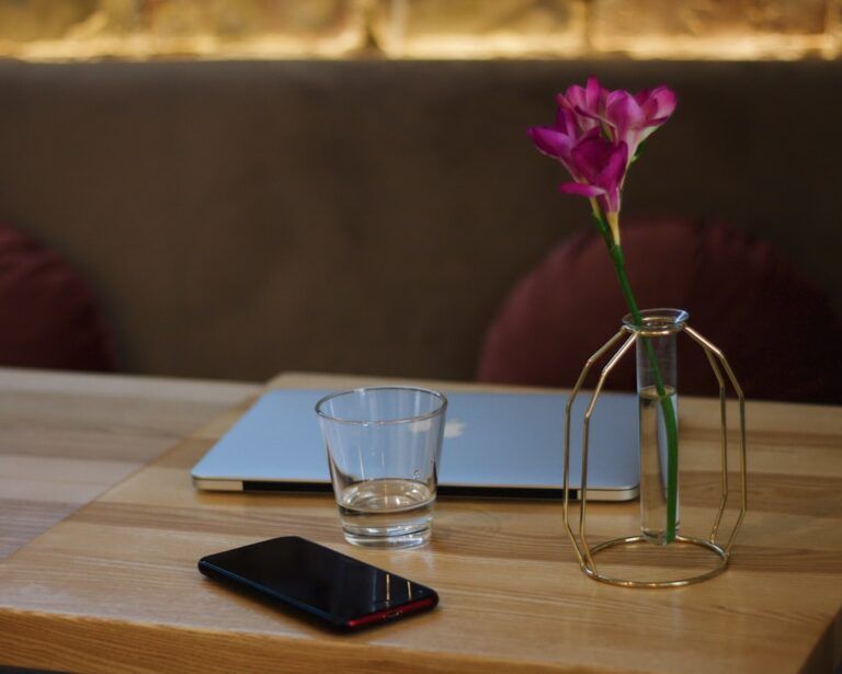 Ein zugeklappter Laptop, ein leeres Glas, ein Smartphone und eine Vase mit einer rosa Blume stehen auf einem Holztisch und spiegeln die Ruhe wider, die man in Aachen oft vorfindet.