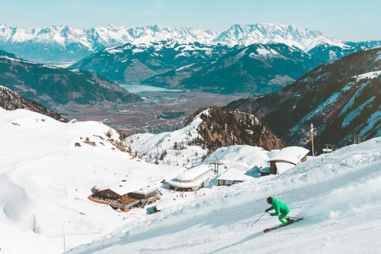 Ein Skifahrer im grünen Anzug fährt einen schneebedeckten Hang hinunter und bietet unter klarem Himmel eine malerische Aussicht auf Berge, ein Tal und einen See in der Ferne, die an die malerischen Landschaften bei Aachen erinnert.